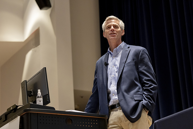Speaker standing near a podium with a computer monitor and microphone, wearing a blazer and dress shirt, in an auditorium setting.